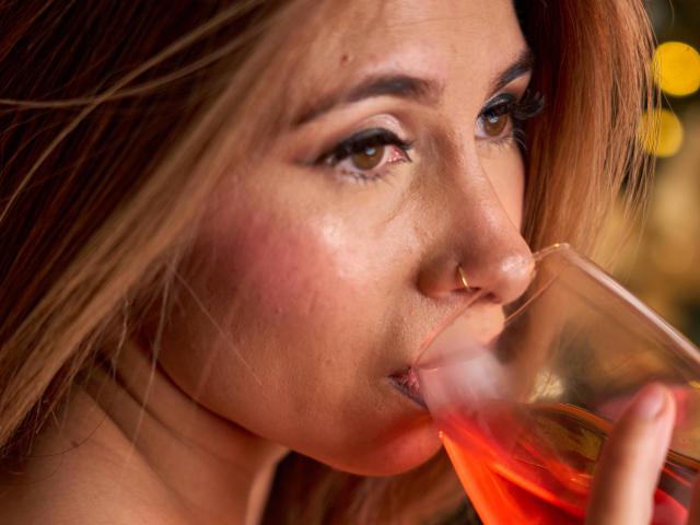 SaraRoades, con un piercing al naso, che beve da un bicchiere di liquido rosso, con le labbra leggermente dischiuse, occhi marroni che guardano verso l'alto, capelli castano chiaro, pelle morbida, trucco leggero, primo piano..