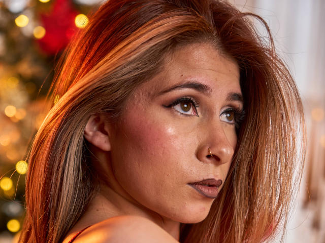 SaraRoades, met bruine ogen, subtiele make-up en roodbruin haar, kijkt over haar schouder, waarbij haar neus en lichtrode wangen zichtbaar zijn. Zonder borsten, geen zichtbare borsten, billen of vagina..