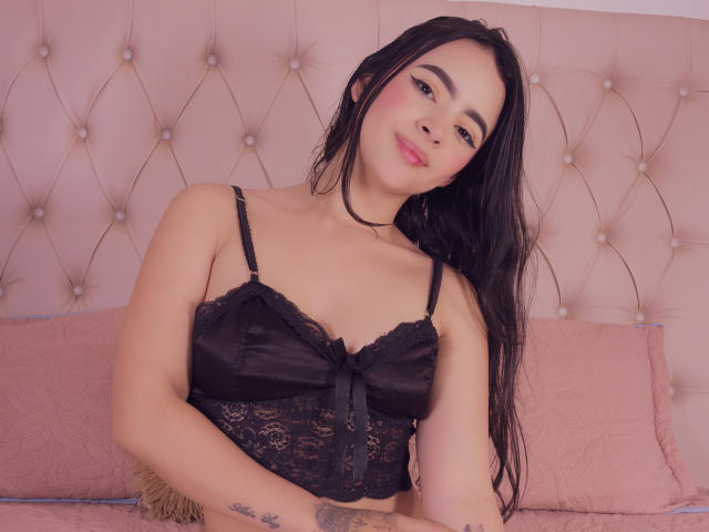 KonanStar with long black hair, wearing a black lace bra, smiling at the camera, showing small tits and a tattoo on her right arm. Pink upholstered headboard in background.