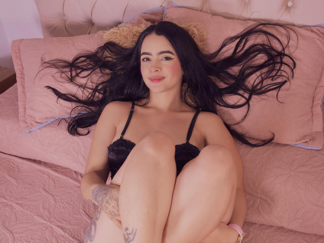 KonanStar with long black hair, black lace bra, and tattoos, lying on a pink bed with legs up, smiling at the camera. Boobs visible, pussy not visible.