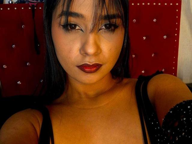 MariamLara with dark hair, red lipstick, and black eyeliner, wearing a black, beaded dress, showing her tits, looking at the camera with a slight, sexy smile.