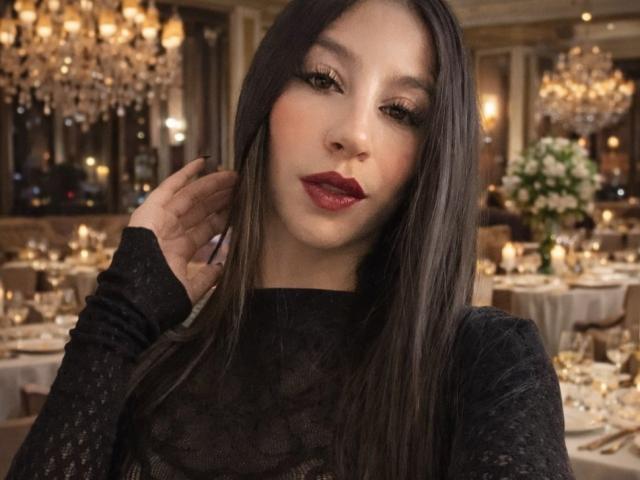 NahomyCarther has long black hair, dark red lipstick, and is wearing a black lace top, with one hand touching her head, in a dimly lit, elegant restaurant.