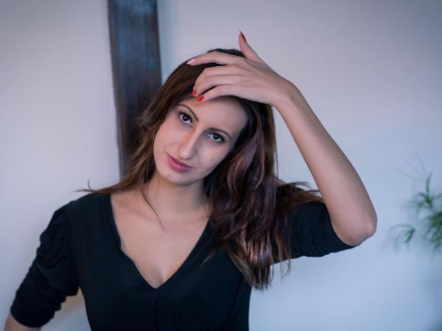 LysAddiction has wavy brown hair, red nails, and a cute face, wearing a black V-neck, with hand on head, looking at camera, showing small tits, in a relaxed pose.
