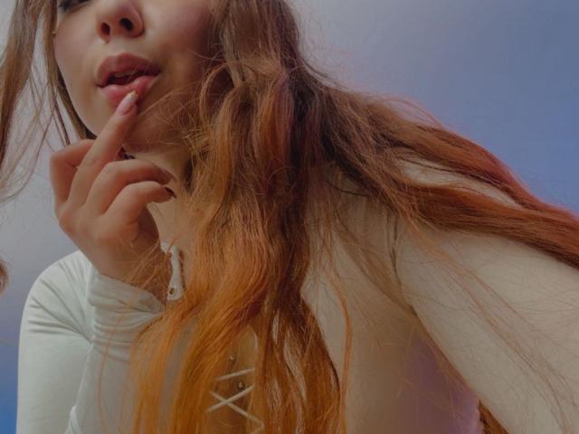 AmberlStha mit langen, orangefarben gefärbten Haaren, einem weißen, schnürbaren Oberteil, einem Finger am Mund, weich-rosa Lippen, leicht sichtbaren Brüsten, einer Hand in der Nähe des Kinns und einem sanften, blau-grauen Hintergrund..