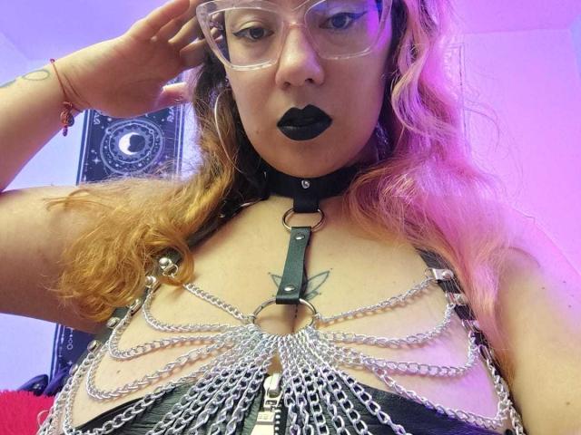 HollyManziniBBW with black lipstick, glasses, and pinkish-brown hair, wearing a metal chain harness over her tits, black leather, and a black choker with a ring.