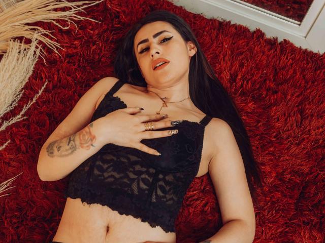 DemiMistral with long black hair, wearing a black lace bra, lying on a red shaggy rug, hand on chest, tattoo on right arm, red lipstick, ring on finger.