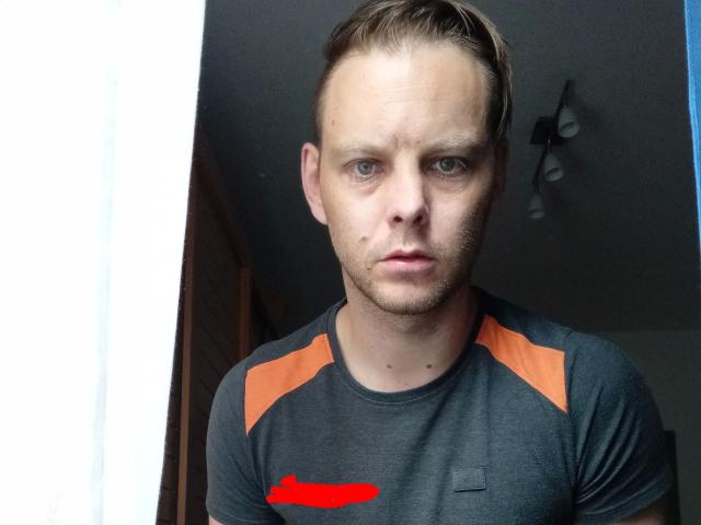 1yayo8-hot is a white man with short, dirty blonde hair and blue eyes. He has a serious, slightly annoyed expression. He's wearing a dark gray t-shirt with orange accents on the shoulders and a red smudge on the chest. The t-shirt has a small, unobtrusive pocket on the left side. The background is blurred, with a wooden door and ceiling light visible. The image is a close-up, focusing on 1yayo8-hot's upper body and face.