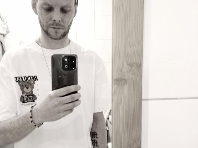 Black and white selfie of 1yayo8-hot with a beard, wearing a white t-shirt with a bear graphic and text, holding a smartphone. 1yayo8-hot has a tattoo on the right arm and wears a bracelet. The t-shirt has a small logo and text. The image is slightly blurred. 1yayo8-hot looks down at the phone, with a neutral expression. The background is mostly white with a wooden panel on the right. Slightly overexposed.