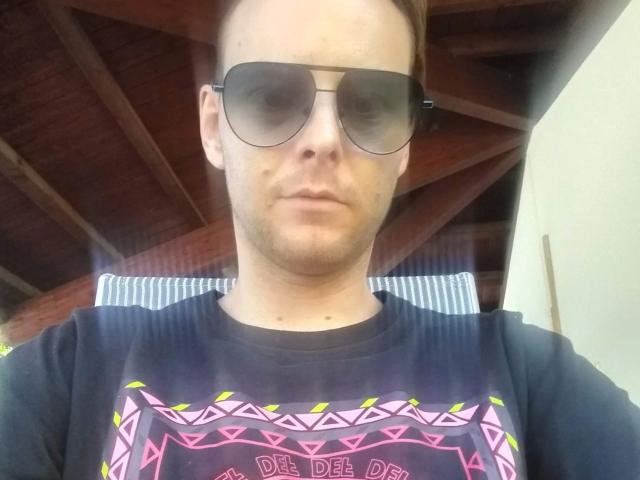 19yayo88-hot indossa occhiali da sole neri e una maglietta nera con motivi tribali rosa e gialli, ha i capelli castani corti e una leggera barba, e guarda direttamente nella fotocamera..