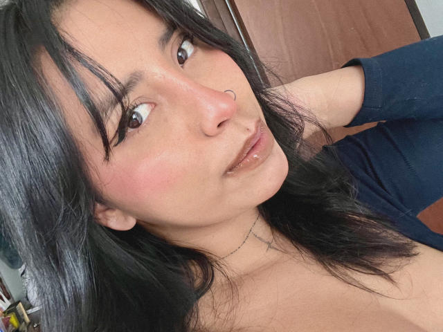 ElizabethMacy has black hair, brown eyes, a nose ring, and glossy pink lips. She's wearing a dark blue shirt, with a slight smile, and her right arm is raised.
