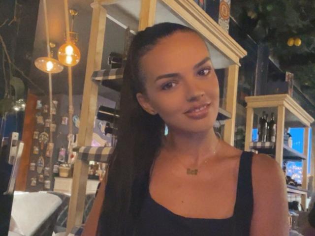 CarolinaPetra with long, slicked-back black hair, wearing a black tank top, smiling subtly, small tits, slim build, and a simple necklace in a warmly lit bar.