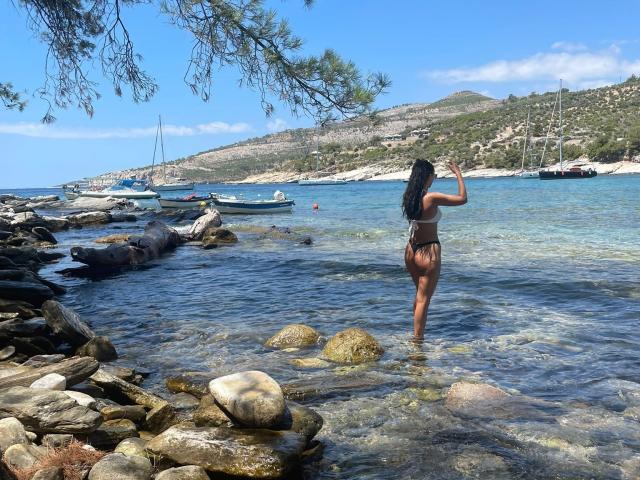 CarolinaPetra with dark skin, long black hair, small brown tits, and a brown thong, standing in clear water, waving, with rocks and boats in the background, under a tree.