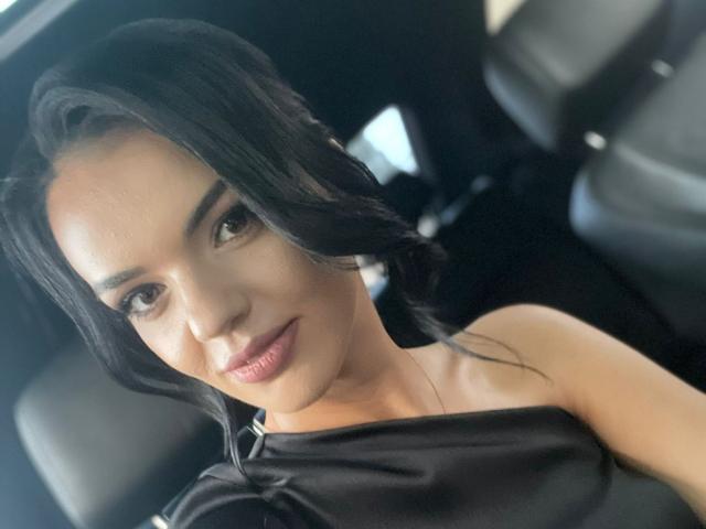 CarolinaPetra with dark wavy hair, brown eyes, and pink lips, wearing a black off-shoulder top, sitting in a car, smiling slightly, showing her bare shoulders and collarbone.