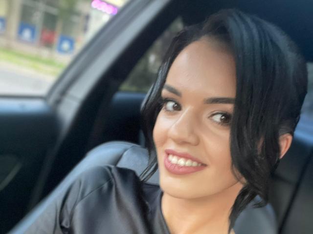 CarolinaPetra with shiny black hair, brown eyes, and pink lipstick, smiling in a car, wearing a black shirt, looking cute and sexy, with a slightly messy hairdo.