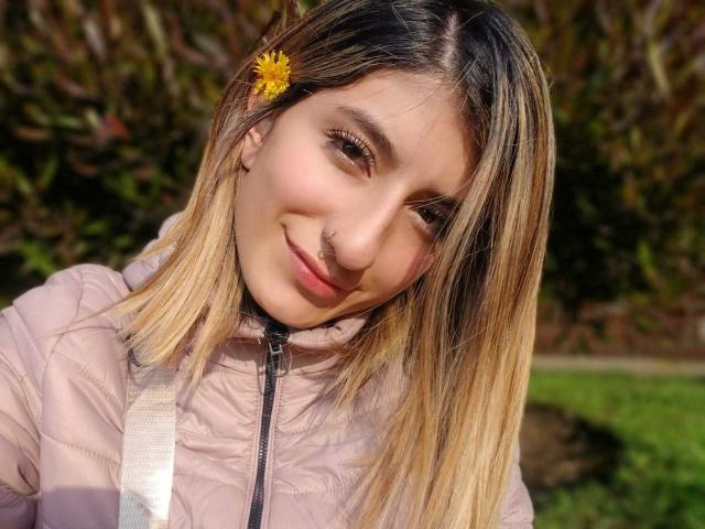 Close-up of Kaiix with light brown hair and a yellow flower in her hair, smiling slightly. She has a small nose ring on her right nostril. Wearing a pink puffer jacket with a zipper. Her skin is fair and she has subtle makeup. The sunlight highlights her hair's blonde tips. The background is blurry with green and brown foliage. The composition focuses on Kaiix's face and upper body. Natural light enhances her features.