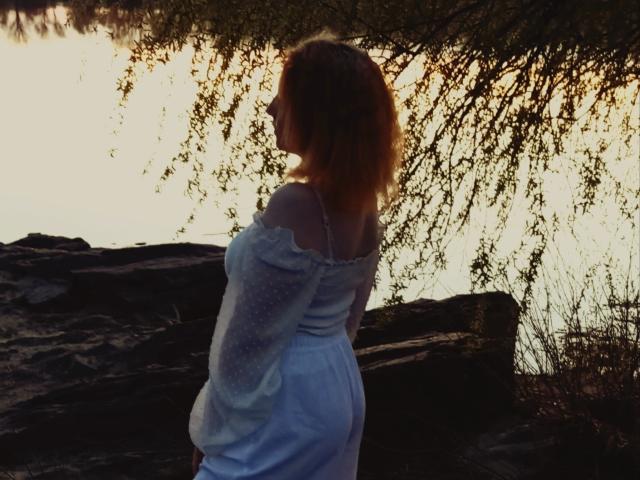 MellMelie stands in profile, facing right, with her back to the camera. She has wavy, shoulder-length red hair. She wears a light blue, off-the-shoulder dress with long sleeves and small polka dots. The dress has a thin strap, visible on her right shoulder. She stands in front of dark rocks and a tree with hanging branches, illuminated by a bright, glowing sunset in the background. The light creates a halo effect around her hair and back. The image uses natural light and has a soft, warm color palette.