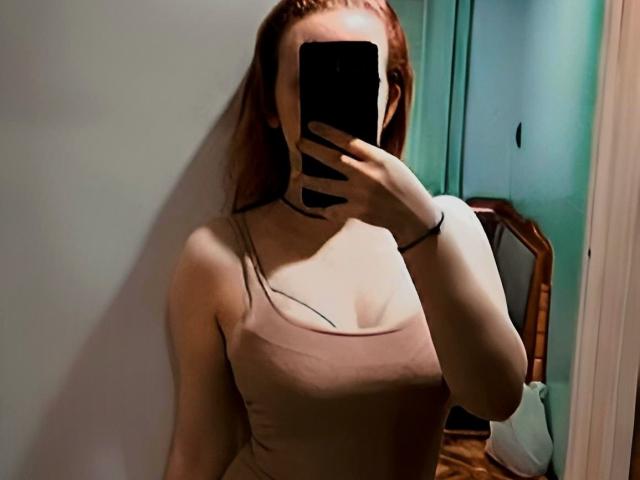 MellMelie mit roten Haaren, die ein Telefon hält, ihr Gesicht bedeckt, ein pinkes Tanktop trägt, ihre Brüste zeigt, in einem Spiegel-Selfie, stehend in einem Raum..
