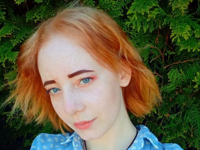 MellMelie has light skin and short, wavy, red-orange hair. Her blue eyes are slightly open, with subtle pink eye shadow on the right side. She has dark, well-defined eyebrows and light pink lips. She wears a blue polka-dot shirt with a black neck strap. The image has a bright, natural light. Her expression is neutral, slightly curious. The texture of her hair is slightly messy. The overall composition is centered and focuses on her face.