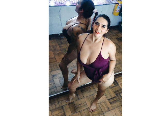 DevhaSexy, con grandes senos, vestida con un traje de baño morado de una sola pieza, de pie frente a un espejo, reflejando su espalda y trasero, sobre un suelo de madera, sonriendo, cabello oscuro, descalza..