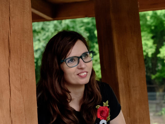 Photograph of SweetLouise. She has long, wavy brown hair and wears black, thick-rimmed glasses. She's smiling slightly, looking to the right. She's dressed in a black t-shirt with a red rose embroidered on the left side. She's standing between wooden pillars. The background is green and blurred, suggesting a forest or park. The wooden pillars are in the foreground, creating a frame around her. Her skin is fair, and she has a casual, approachable look. The image is clear and well-lit.