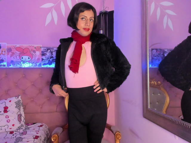 EimyFlorian has short black hair, wearing a black fur jacket, red scarf, pink top, and black pants, standing with hands on hips, in a pink room.