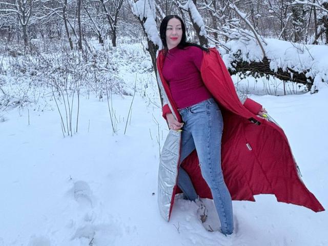 MirandaHott69 with red coat, pink top, blue jeans, standing in snow, holding white backpack, leaning against snow-covered tree, black hair, red lipstick, snowy forest background.