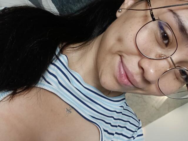 Karollteyes, con cabello negro, usando gafas redondas, sombra de ojos morada y una camiseta sin mangas a rayas, mostrando un piercing en la nariz y un pequeño tatuaje de una cruz en su hombro derecho..