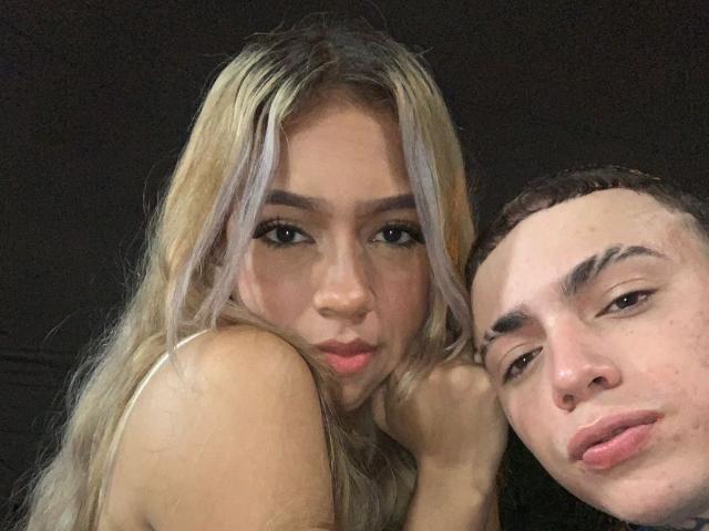 IceCumHot with long blonde and purple-tipped hair, pouty pink lips, and small tits, next to a bald, brown-skinned guy with thick eyebrows and puffy lips, both in selfie.