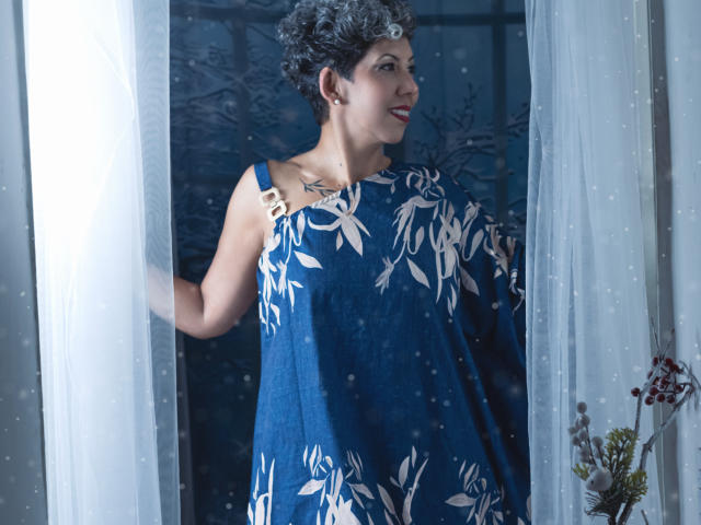 JuliaNewell with short curly gray hair, blue dress with white leaf pattern, standing in doorway, white curtains, holding left curtain, small tattoo on right side of neck, smiling.