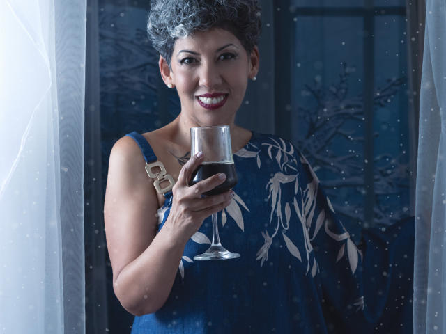 JuliaNewell with short curly gray hair, wearing a blue dress with leaf patterns, holding a glass of red wine, smiling, standing by a window with white curtains.