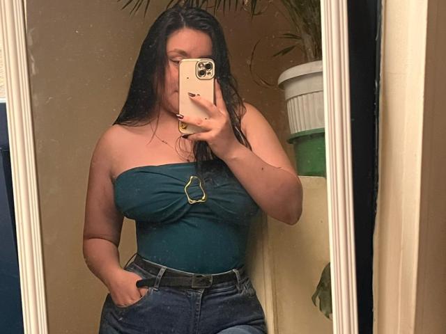 EmaJames with wet black hair, wearing a green strapless top, showing her boobs, and blue jeans, taking a mirror selfie with her phone, hand in pocket.