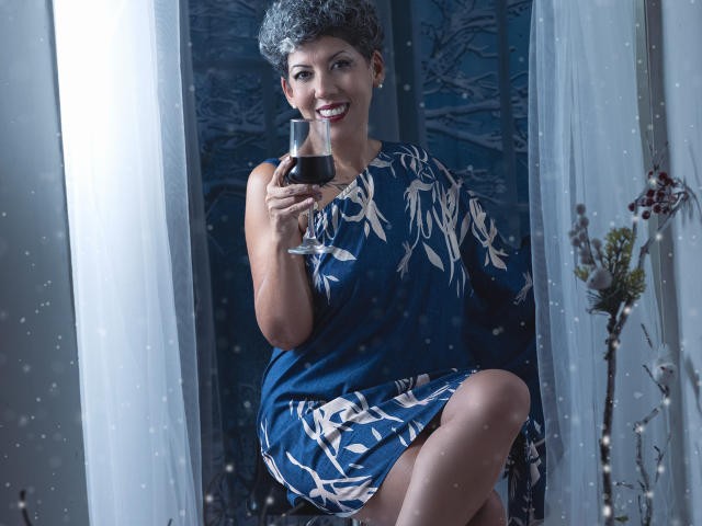 JuliaNewell with short curly gray hair, wearing a blue dress with white leaf patterns, holding a glass of red wine, smiling, sitting with one leg raised, white curtains.