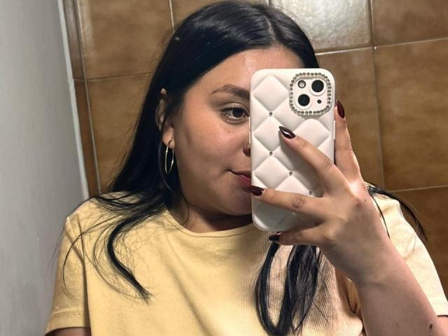 EmaJames with long black hair, wearing a yellow shirt, holding a white phone with diamond-patterned case, taking a mirror selfie, showing her red-painted nails and hoop earrings.
