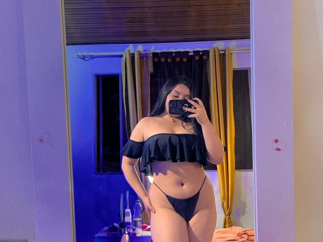EmaJames, com longos cabelos pretos, vestindo uma blusa preta com um ombro descoberto e uma tanga preta, está em frente a um espelho, segurando um celular, mostrando seus seios e sua vulva..