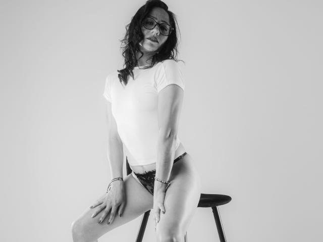 SamanthaCorine with wavy dark hair, glasses, white t-shirt, and black lace panties, sitting on a stool, legs apart, hands on thighs, smiling. Monochrome photo.