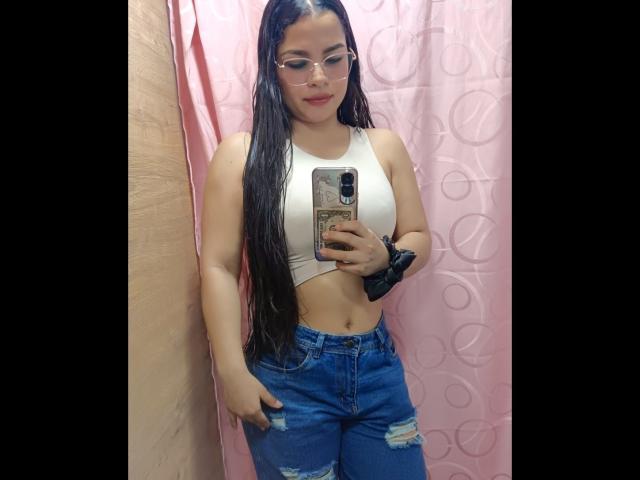 AmaiaSaenz with long black wet hair, glasses, white crop top, blue ripped jeans, black scrunchie, holding phone, pink curtain, showing her tits, standing in a bathroom.