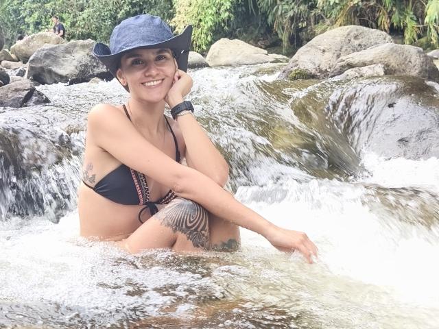 AmelieLautrec, wearing a black bikini and blue hat, sits in a rocky stream, smiling with hand on cheek, showing tattoos on thigh and arm, with water splashing.