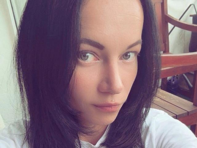 Dirtyheike-hot has black hair, green eyes, a small nose piercing, and a serious expression. She's wearing a white shirt, with a wooden chair in the background.