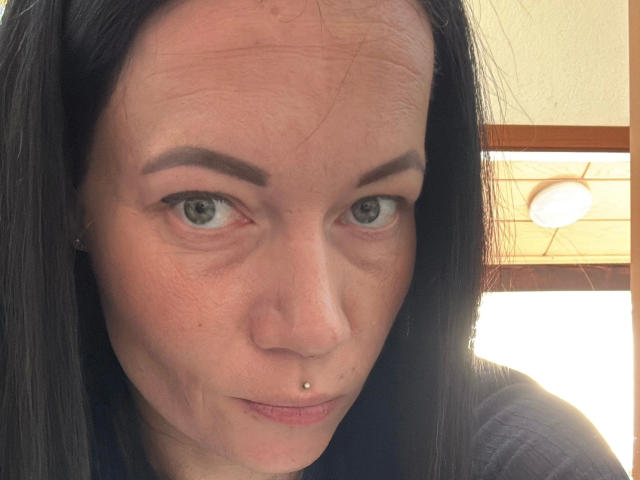Dirtyheike-hot has pale skin, black straight hair, green eyes, and a pierced lower lip. She's looking at the camera with a slightly serious expression.