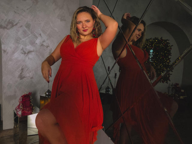 SallyVerda with blonde hair, red dress, and red earrings, standing with one leg raised, arm raised, reflecting in a mirror, showing her red dress and tits.