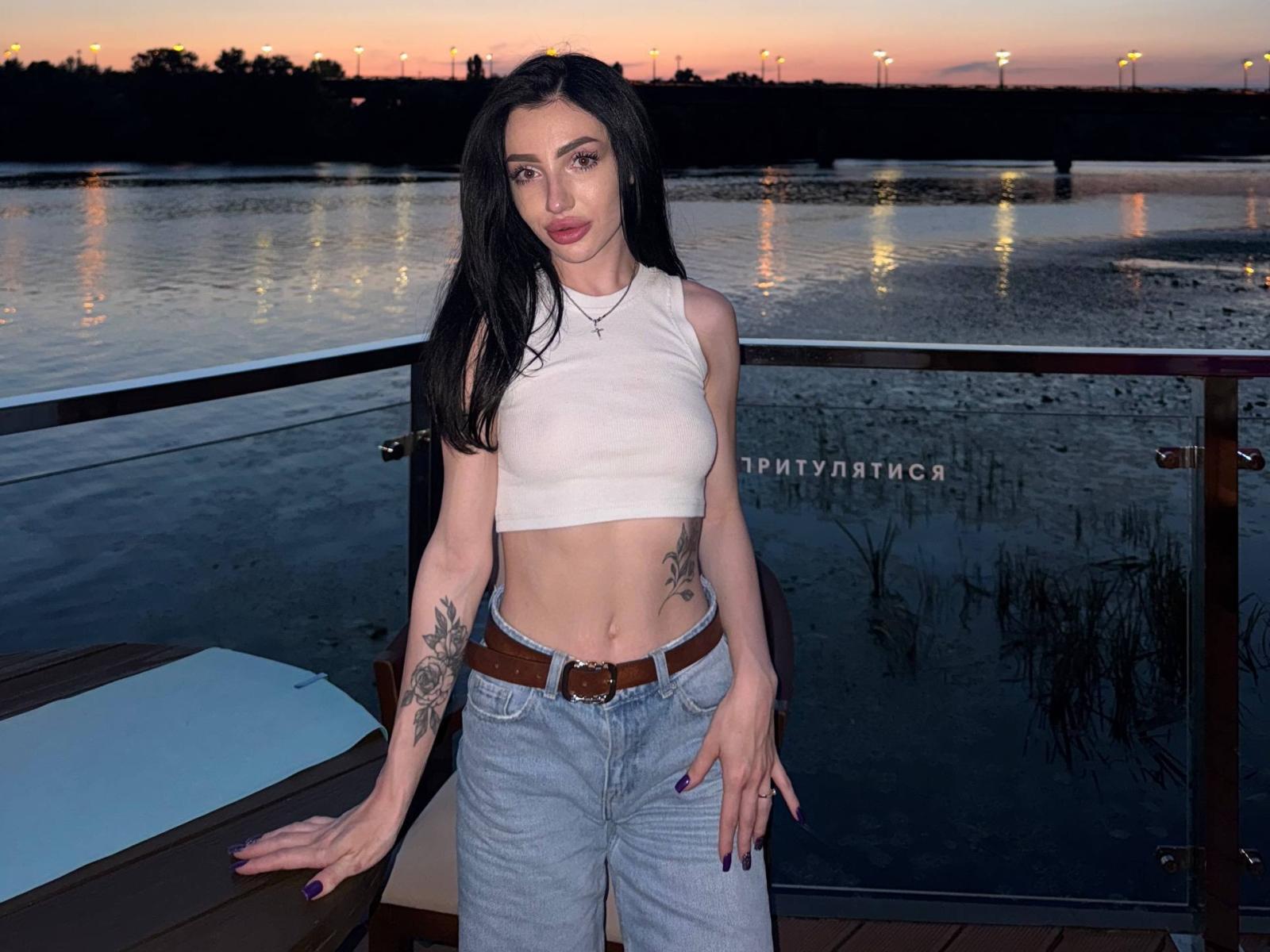 NancyLovvee, com cabelos longos e pretos, blusa cropped branca, tatuagens nos braços, jeans azuis, cinto marrom, em pé perto de um parapeito de vidro ao pôr do sol, com as mãos em uma mesa de madeira, reflexo da água..