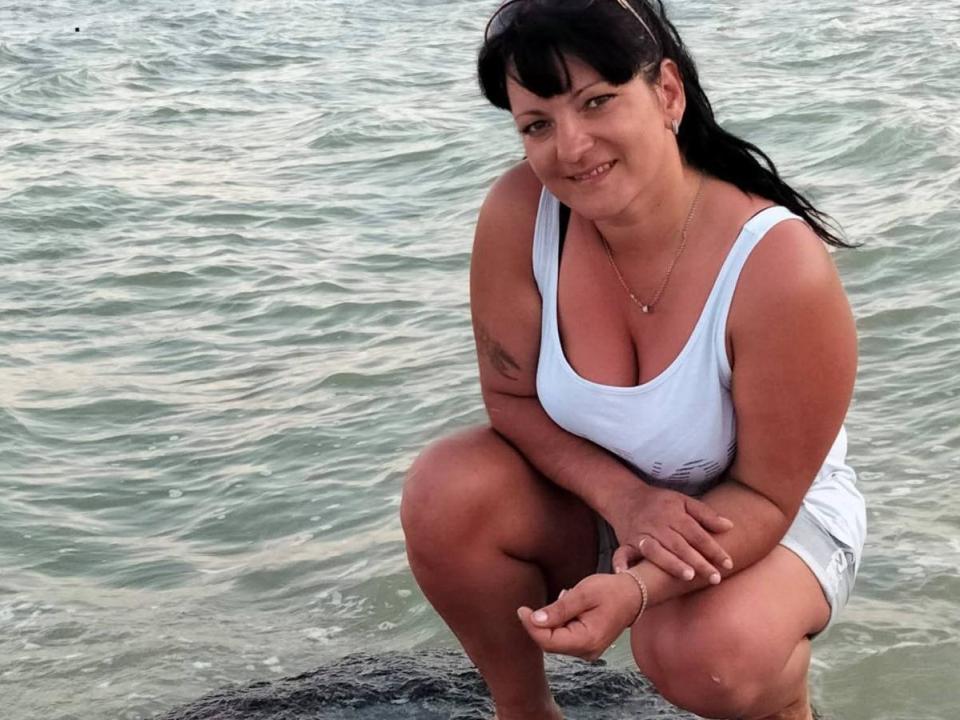 CoraBlack, une femme corpulente avec des cheveux noirs et une frange, est accroupie sur un rocher, vêtue d'un débardeur blanc qui met en valeur ses seins volumineux, et elle sourit à l'appareil photo..