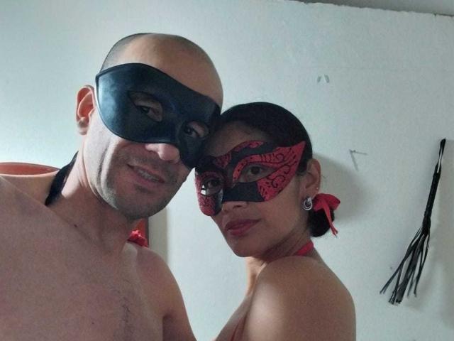 A photograph of CoupleHot6s. A bald, light-skinned man with a slight smile wears a black mask. Next to him, a woman with dark hair in a bun, wearing a red mask with black lace patterns, and red ribbon in her hair. Both are topless, with the man's shoulder visible. The woman's right eye is exposed. A black flogger is in the background on the right. The man's face is slightly to the left, and the woman's face is to the right.