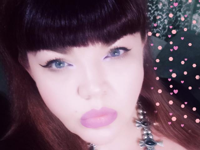 Close-up of RedBustyX with pale skin, blue eyes, and dark brown, straight hair with blunt bangs. She has full, pink lips and wears black eyeliner. Her necklace has a silver, floral design. Pink heart emojis are digitally added to the right side. Her expression is neutral, slightly pouty. The image has a soft, almost pastel-like color palette. The focus is on her face, with the background blurred.