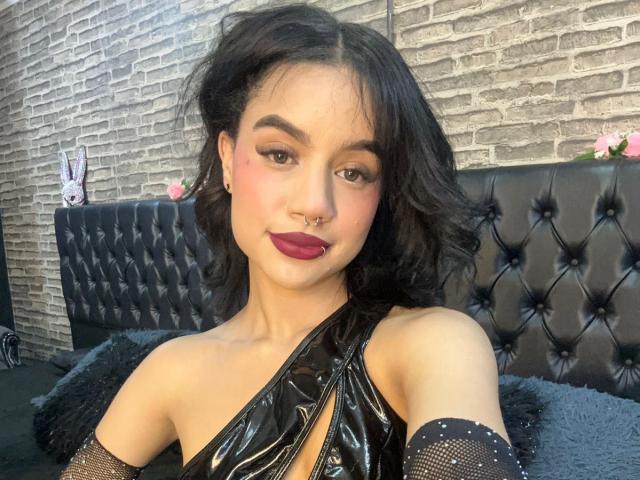 WendyJames with dark wavy hair, black vinyl top, nose ring, pink eye makeup, red lipstick, black mesh sleeves, looking at camera, sitting on black tufted bed.