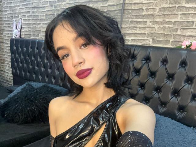 WendyJames with black wavy hair, nose ring, and red lipstick, wearing a shiny black one-shoulder top, sitting on a black tufted bed, showing her bare shoulder and upper chest.