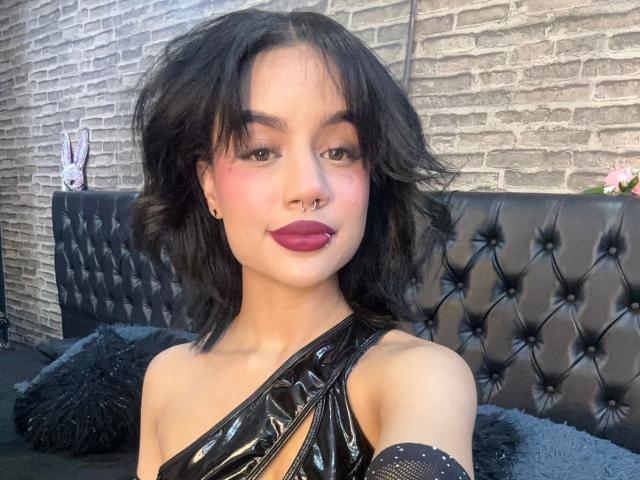 WendyJames with black wavy hair, nose ring, and dark lipstick, wearing a shiny black one-shoulder top, showing her tits, sitting on a black tufted leather couch.
