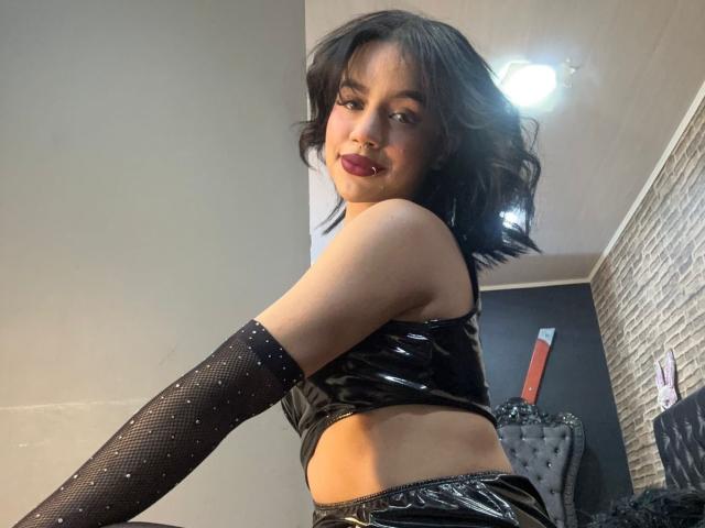 WendyJames with black wavy hair, dark lipstick, and a shiny black crop top, wearing black sparkly arm sleeves, looking over her shoulder, showing her bare midriff and ass.