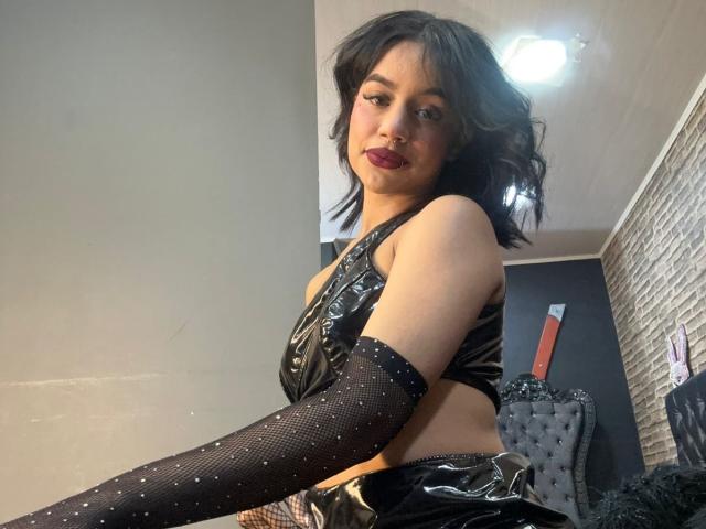WendyJames with black wavy hair, wearing a shiny black leather top, matching arm warmers, and red lipstick, sitting in a room, showing her bare midriff and small ass.
