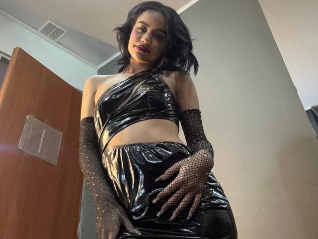 WendyJames with black shiny latex outfit, one-shoulder top, high-waisted skirt, and black fishnet gloves. She has wavy black hair and is looking down at the camera.